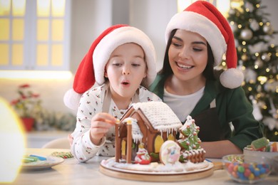 Photo of Mother and daughter decorating gingerbread house at table indoors
