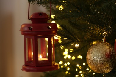 Photo of Christmas lantern with burning candle, festive lights and ball on fir tree against blurred background, closeup
