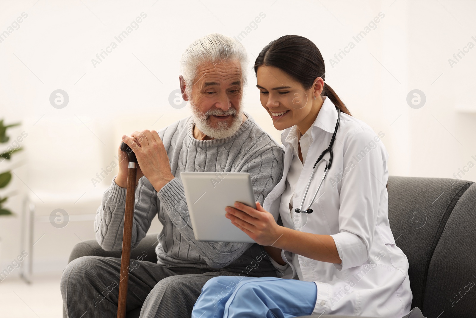 Photo of Smiling nurse with tablet and elderly patient on sofa in hospital