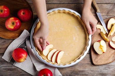 Photo of Woman putting apple slice into baking dish with dough to make traditional English pie at wooden table, top view