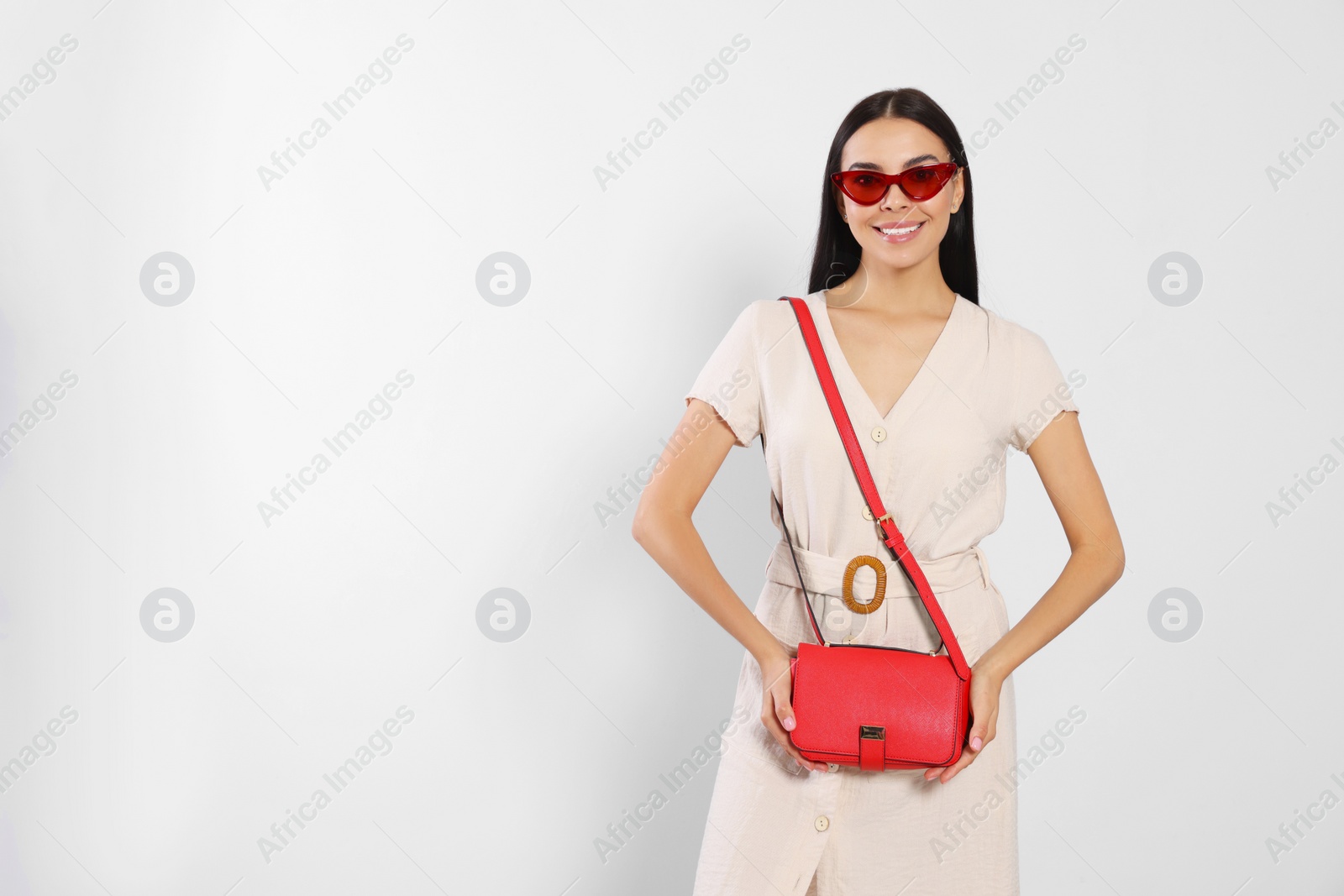 Photo of Beautiful young woman with red stylish bag in sunglasses on light grey background, space for text