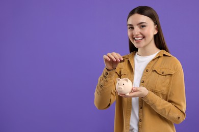 Photo of Happy woman putting coin into piggy bank on purple background, space for text