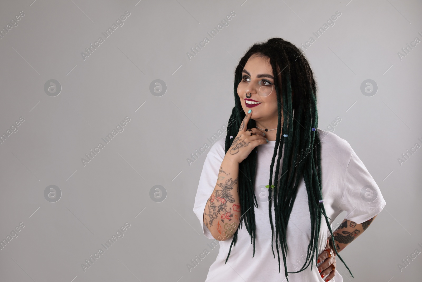 Photo of Beautiful young woman with tattoos on arms, nose piercing and dreadlocks against grey background. Space for text