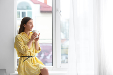 Photo of Young woman with cup on window sill indoors
