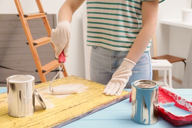Woman using roller to paint plank with white dye at light blue wooden table indoors, closeup
