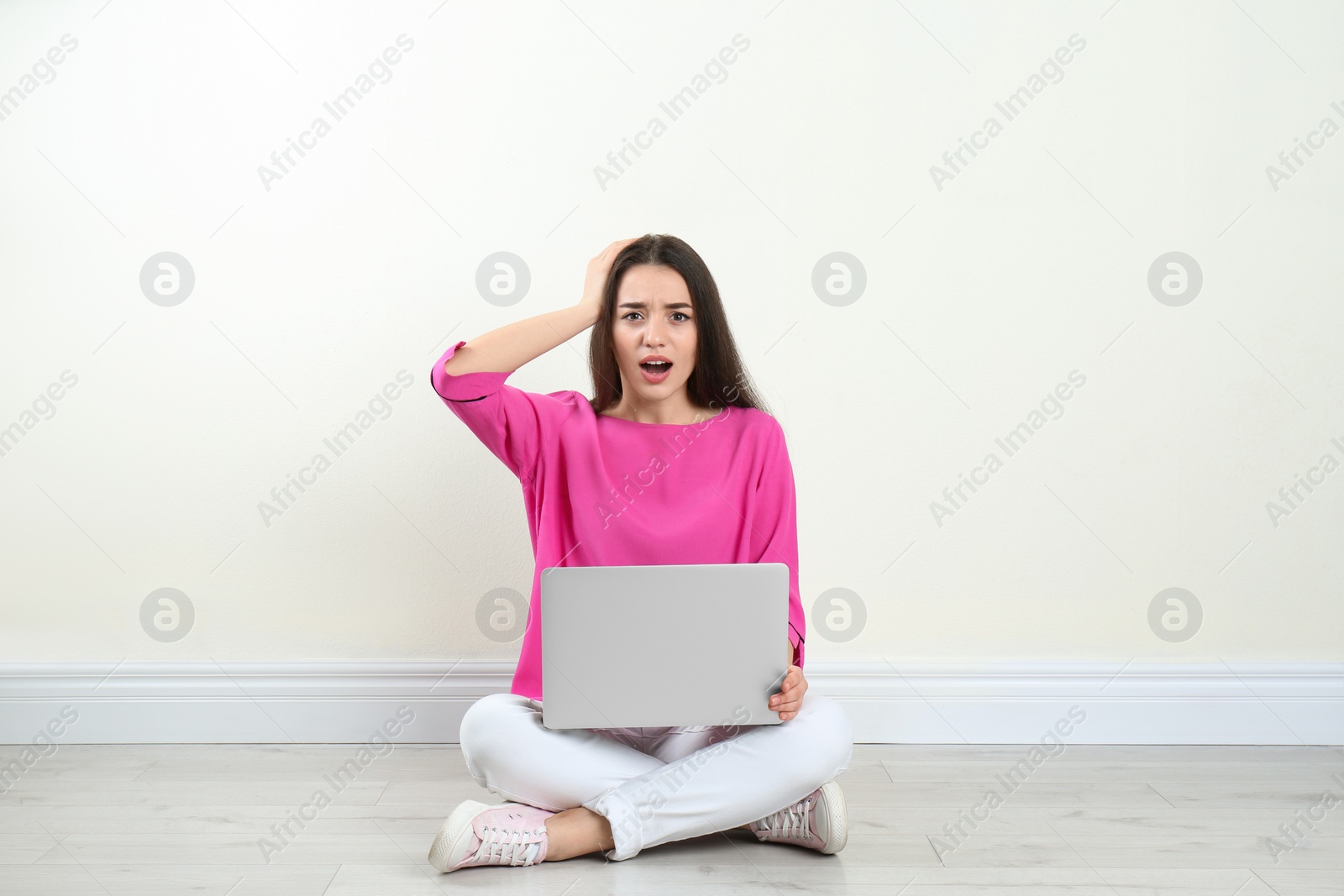 Photo of Shocked young woman with laptop sitting on floor near light wall indoors