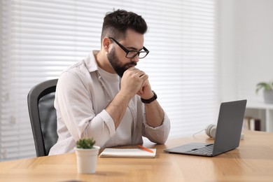 Photo of Young man in glasses watching webinar at table in room