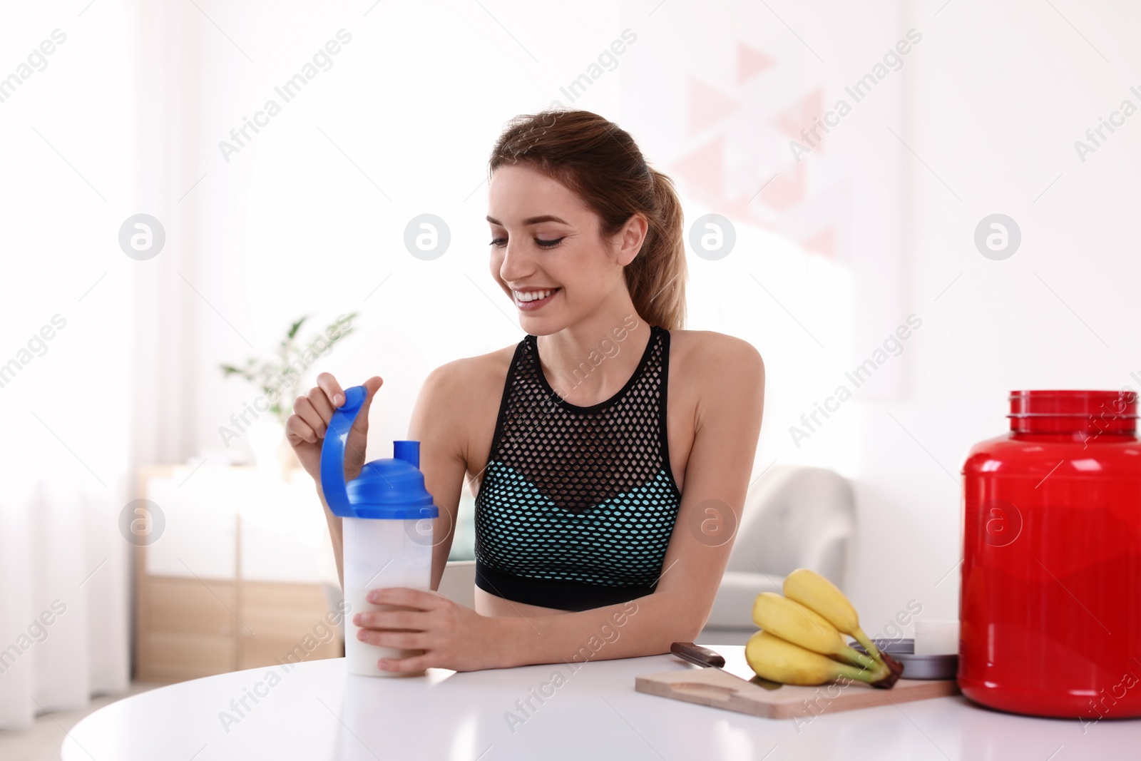 Photo of Young woman holding bottle of protein shake at table with ingredients in room