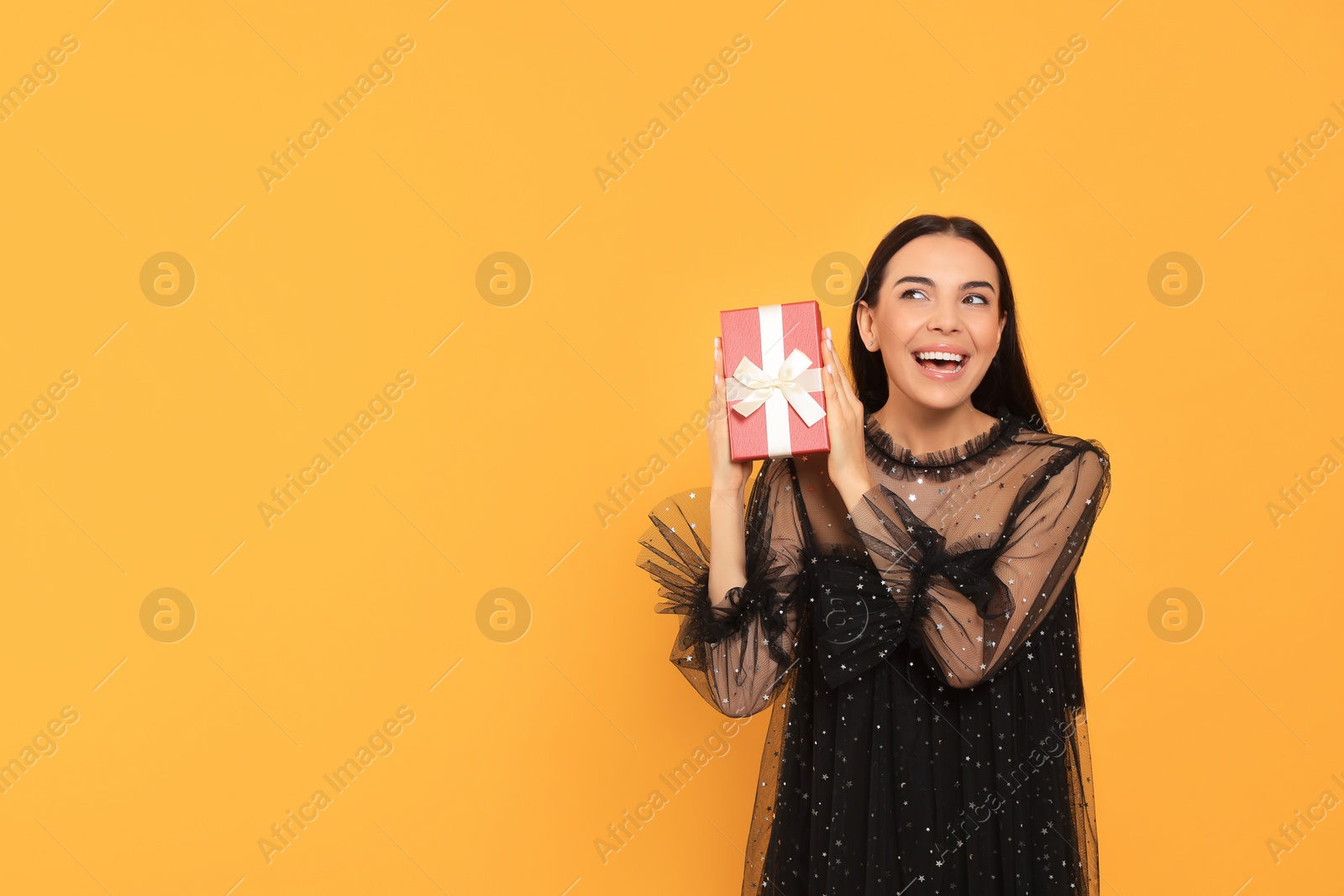 Photo of Emotional young woman in festive dress with gift box on orange background, space for text
