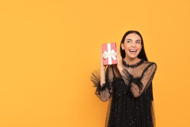 Photo of Emotional young woman in festive dress with gift box on orange background, space for text