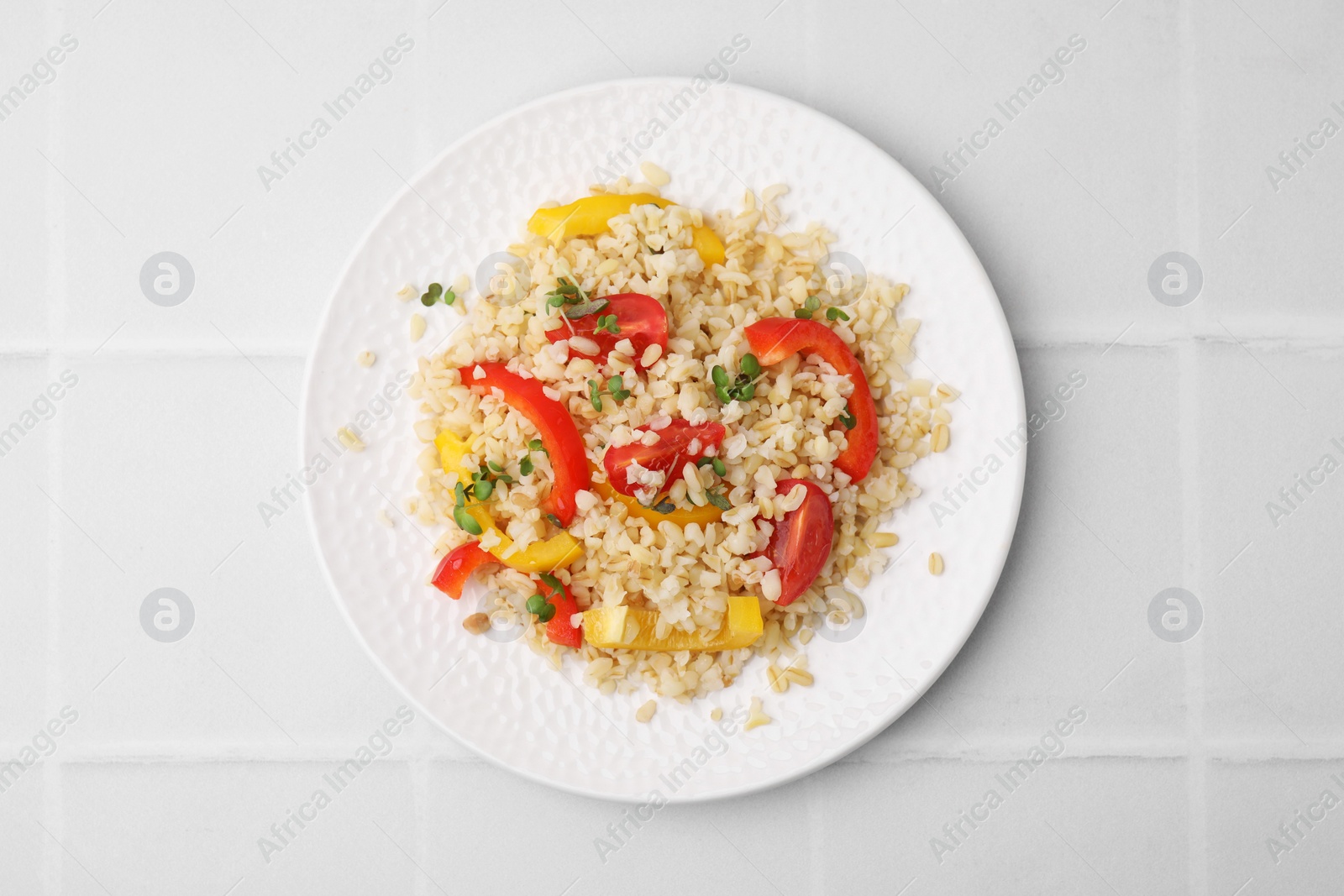 Photo of Plate of cooked bulgur with vegetables on white tiled table, top view