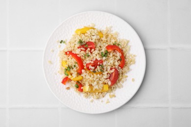 Plate of cooked bulgur with vegetables on white tiled table, top view