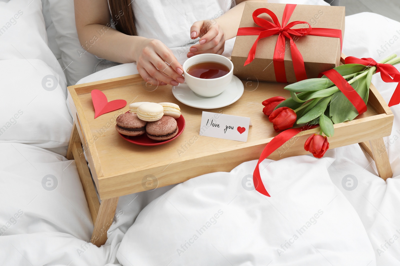 Photo of Tasty breakfast served in bed. Woman with tea, macarons, gift box, flowers and I Love You card at home, closeup