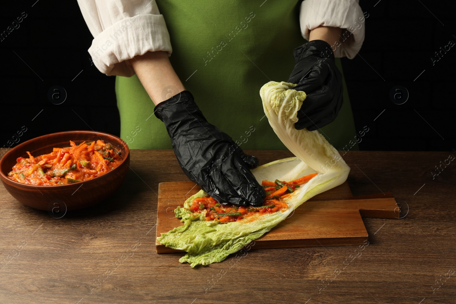 Photo of Woman preparing spicy cabbage kimchi at wooden table against dark background, closeup