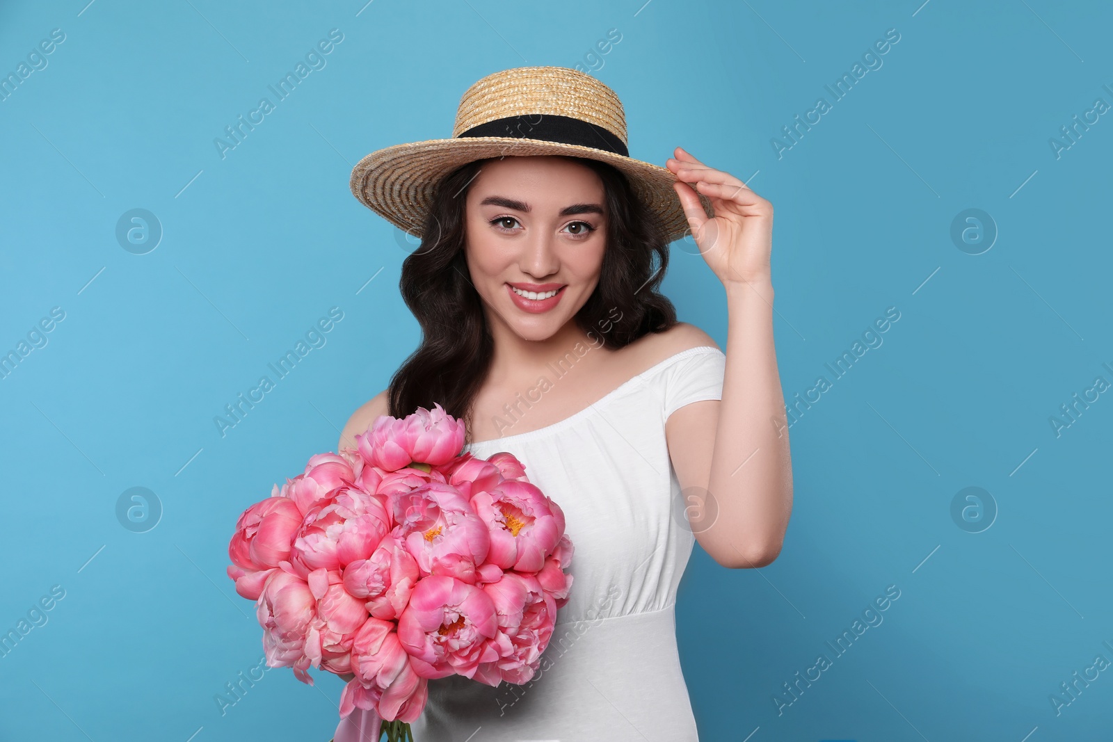 Photo of Beautiful young woman in straw hat with bouquet of pink peonies against light blue background