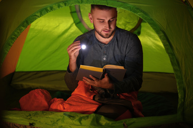 Young man with flashlight reading book in tent