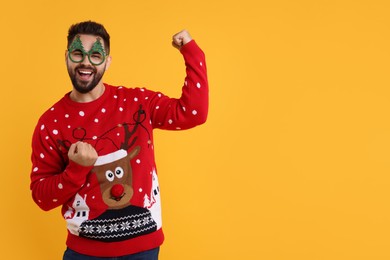 Photo of Happy young man in Christmas sweater and funny glasses on orange background. Space for text