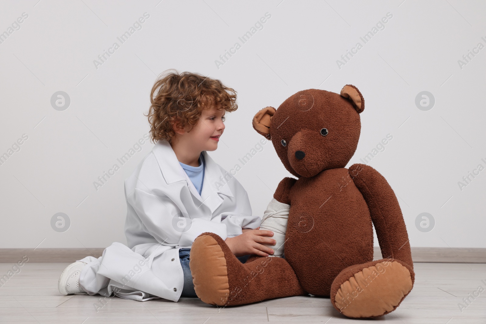 Photo of Little boy in medical uniform and toy bear with bandage indoors