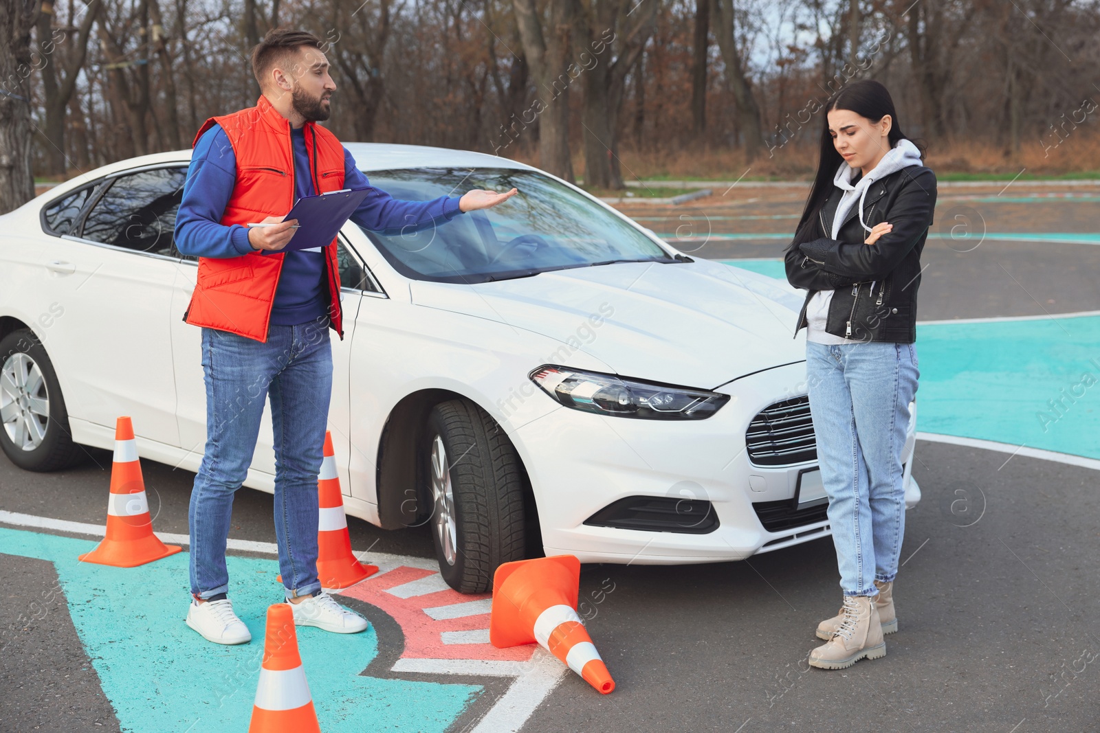 Photo of Young woman with instructor near car and fallen traffic cone outdoors. Failed driving school exam