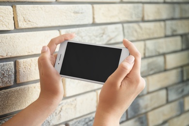 Photo of Young woman holding mobile phone with blank screen in hands near brick wall