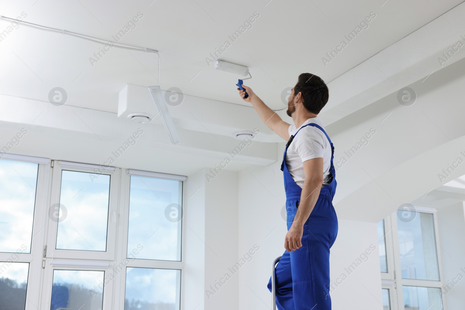 Photo of Handyman with roller painting ceiling on step ladder in room