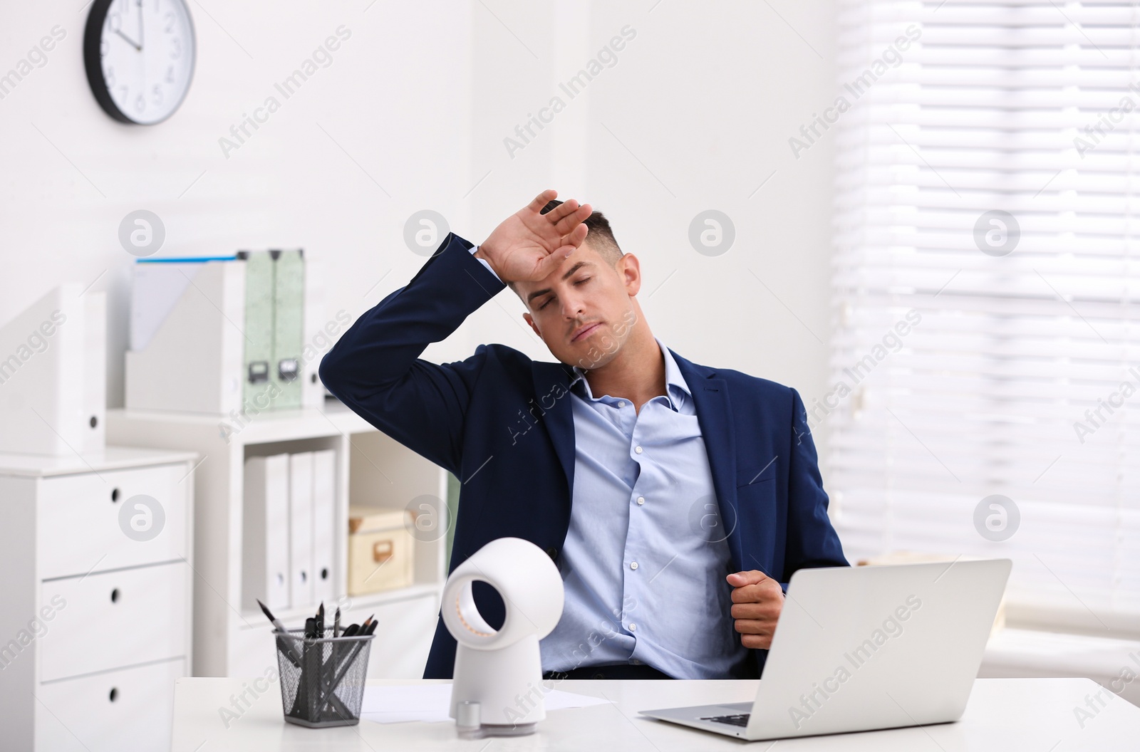 Photo of Man suffering from heat in front of fan at workplace