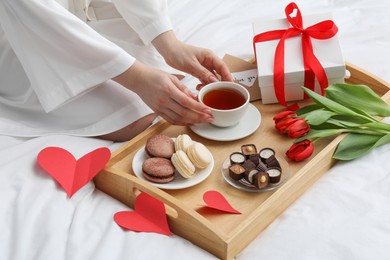 Photo of Tasty breakfast served in bed. Woman with tea, desserts, gift box and flowers at home, closeup