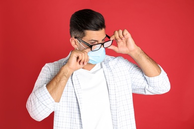 Man wiping foggy glasses caused by wearing medical mask on red background