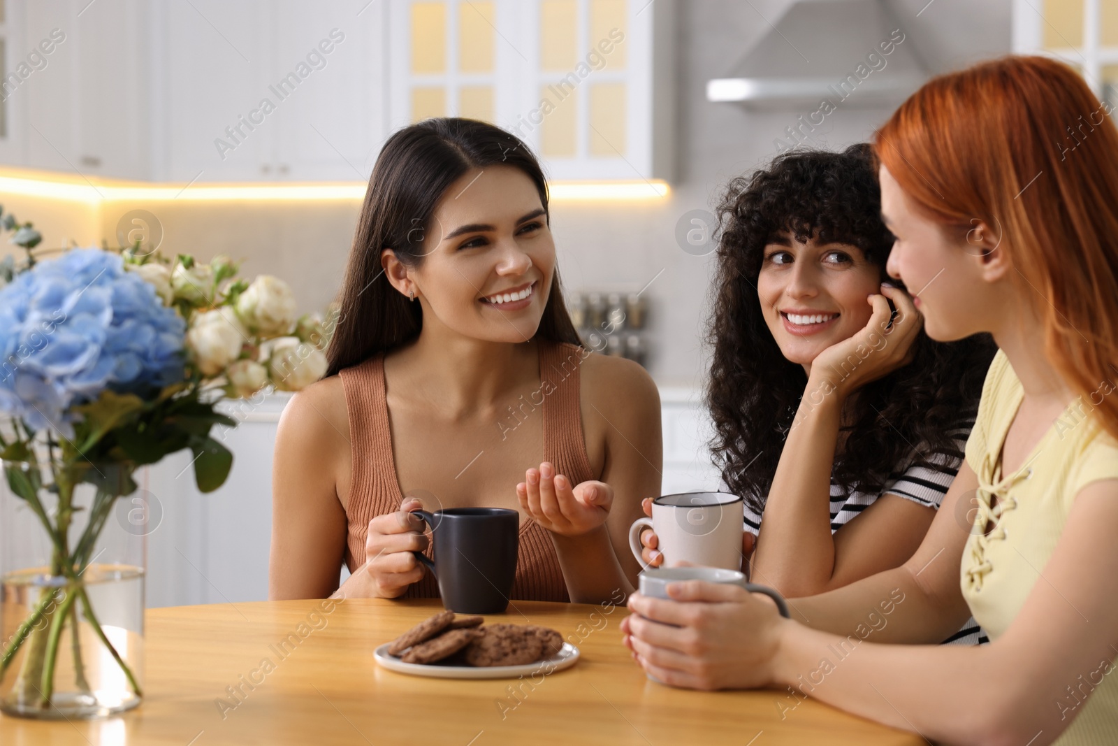 Photo of Happy young friends with cups of drink spending time together at table in kitchen