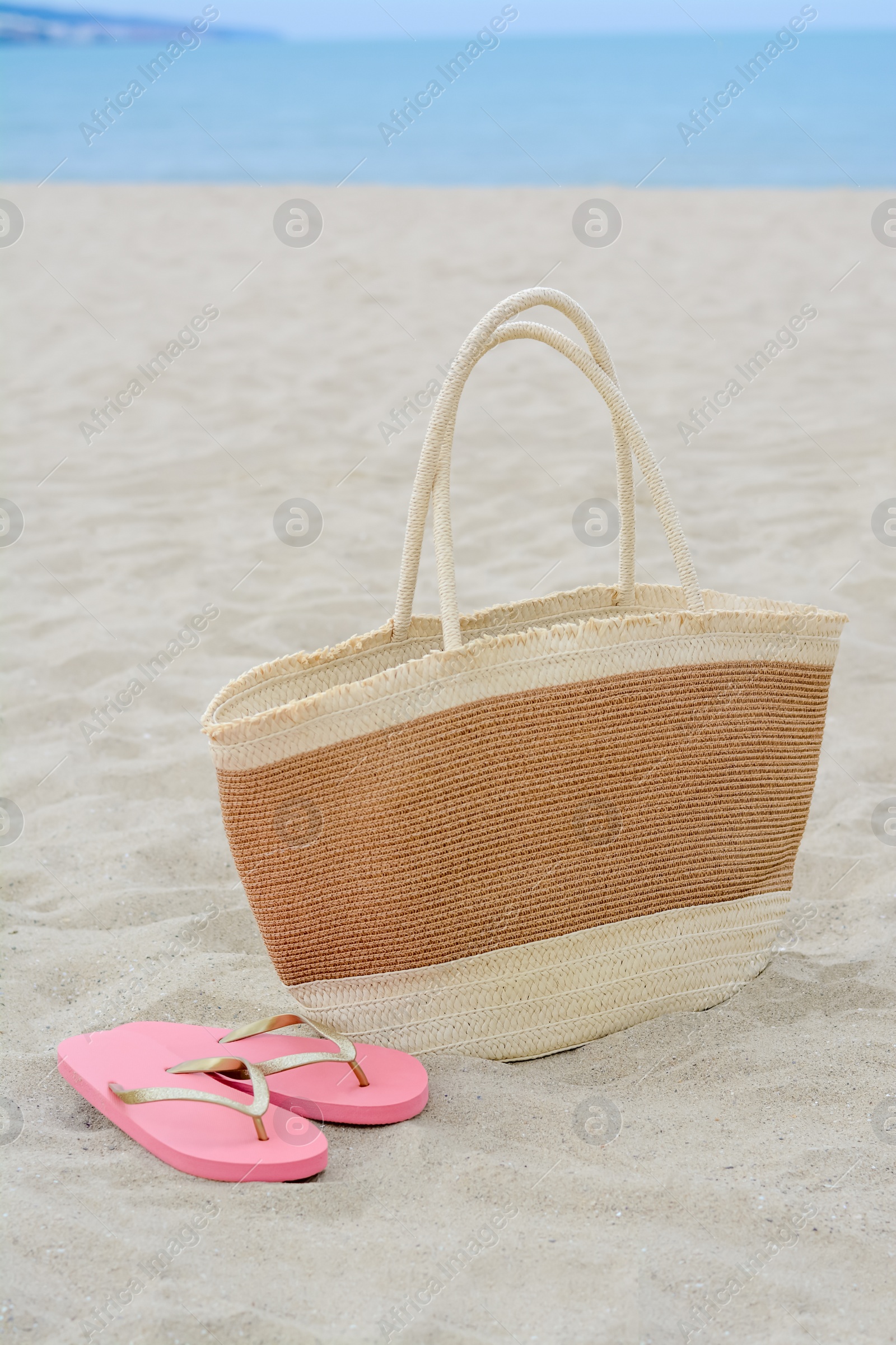 Photo of Stylish straw bag and flip flops on sand near sea. Beach accessories