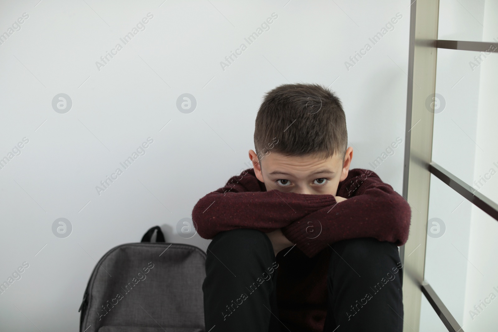 Photo of Upset boy with backpack sitting near wall indoors. Space for text