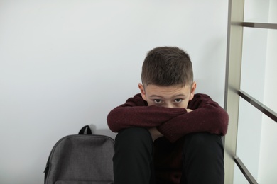 Upset boy with backpack sitting near wall indoors. Space for text