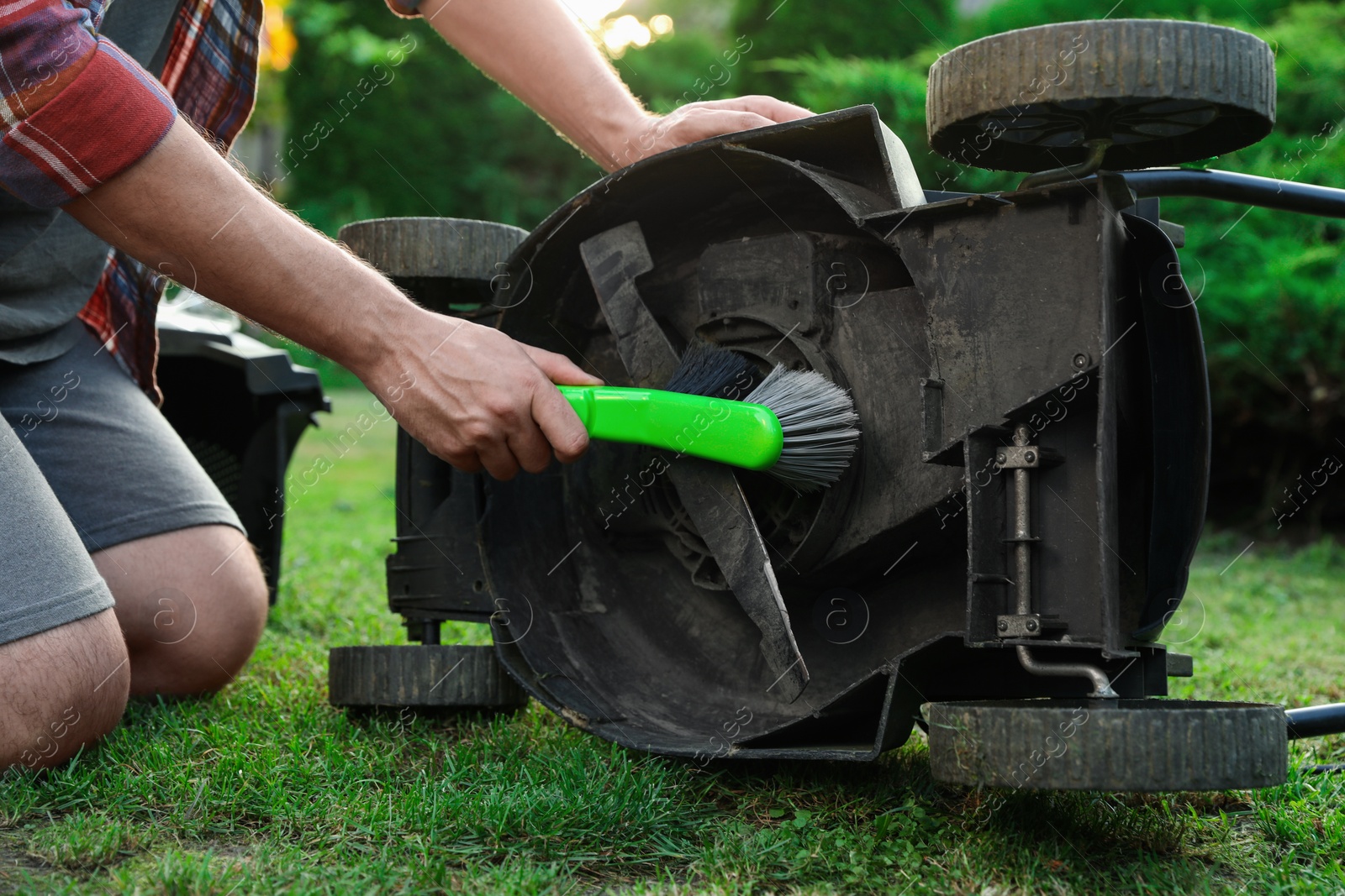 Photo of Man cleaning lawn mower with brush in garden, closeup