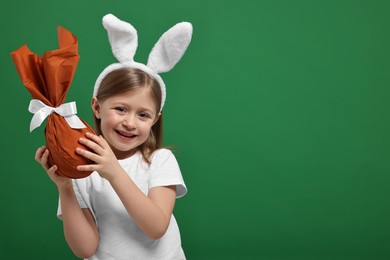 Photo of Easter celebration. Cute girl with bunny ears holding wrapped gift on green background, space for text