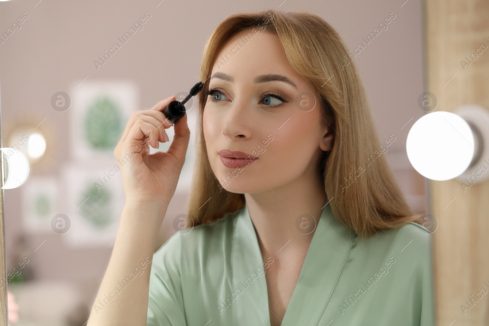 Photo of Reflection of beautiful young woman applying mascara in mirror at home