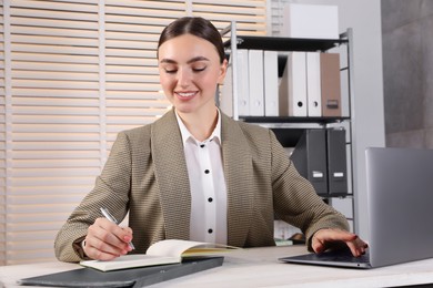 Photo of Happy woman taking notes while using laptop at light wooden table in office