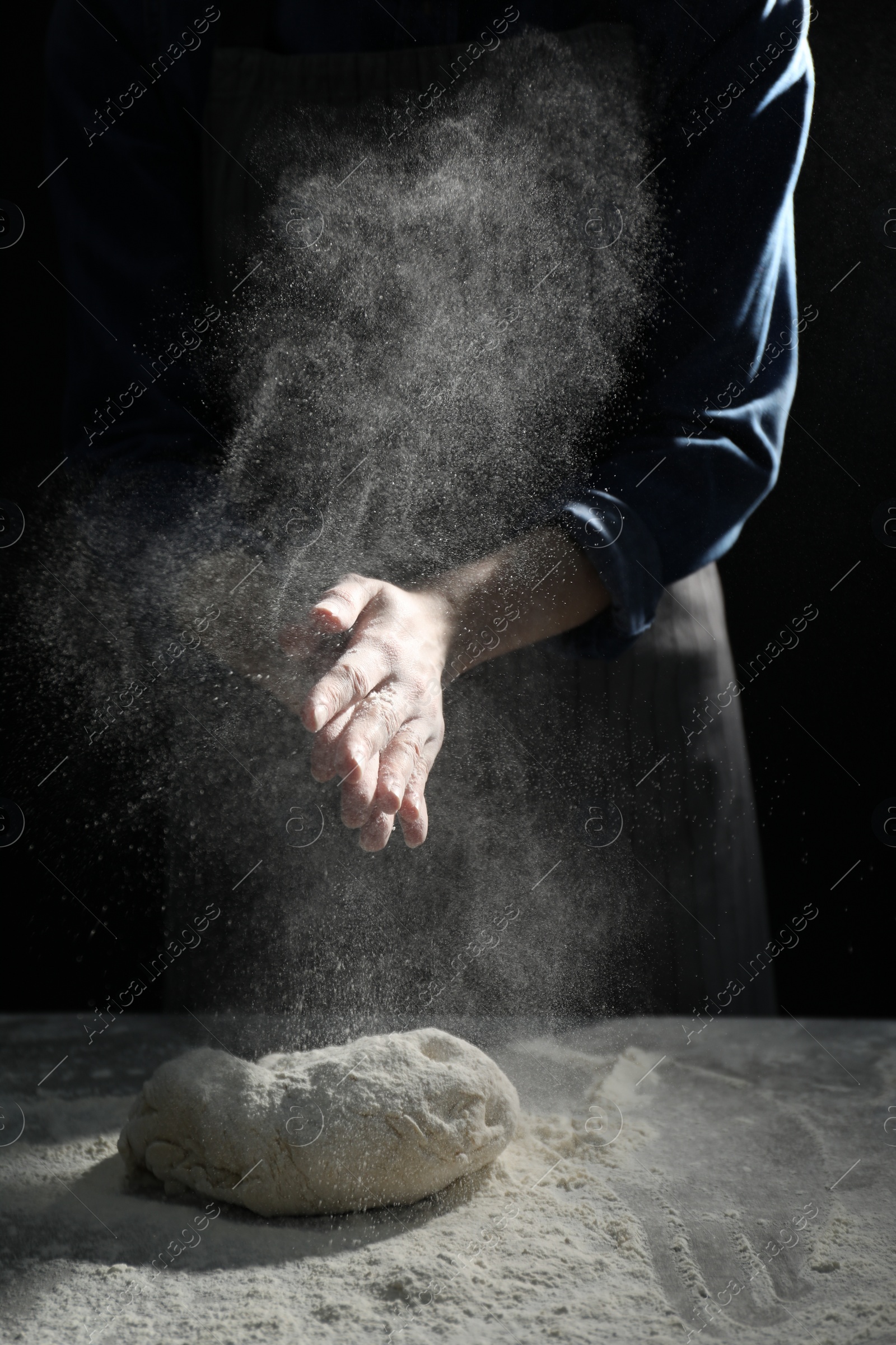 Photo of Making bread. Woman sprinkling flour over dough at table on dark background, closeup