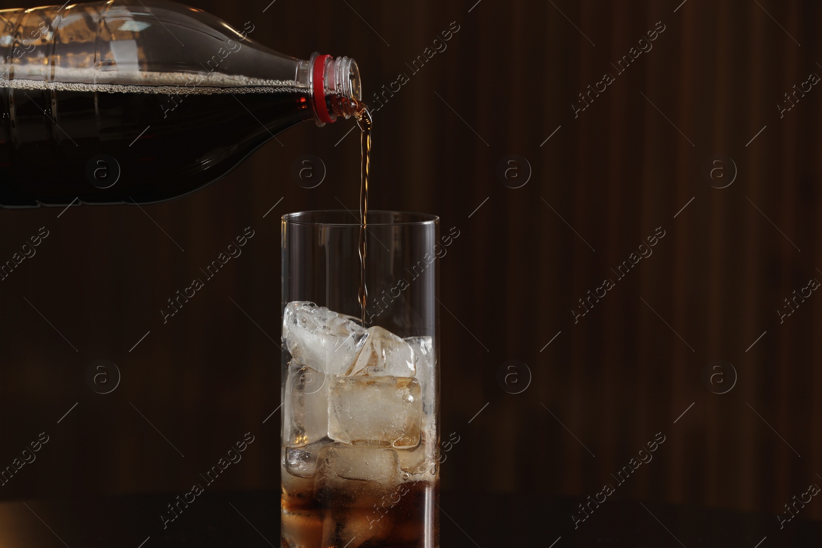 Photo of Pouring cola from bottle into glass with ice cubes on table against blurred background. Space for text