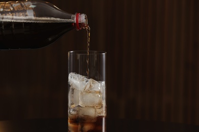 Photo of Pouring cola from bottle into glass with ice cubes on table against blurred background. Space for text