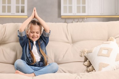 Little girl meditating on soft sofa at home, space for text