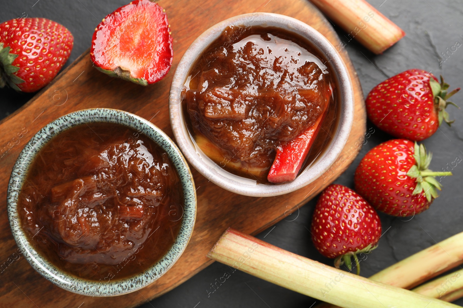 Photo of Tasty rhubarb jam in bowls, stems and strawberries on dark table, flat lay