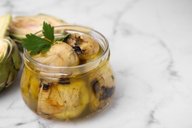 Photo of Jar of delicious artichokes pickled in olive oil and fresh vegetables on white marble table, closeup. Space for text