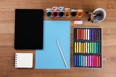 Photo of Blank sheet of paper, colorful chalk pastels, tablet and other drawing tools on wooden table, flat lay. Modern artist's workplace