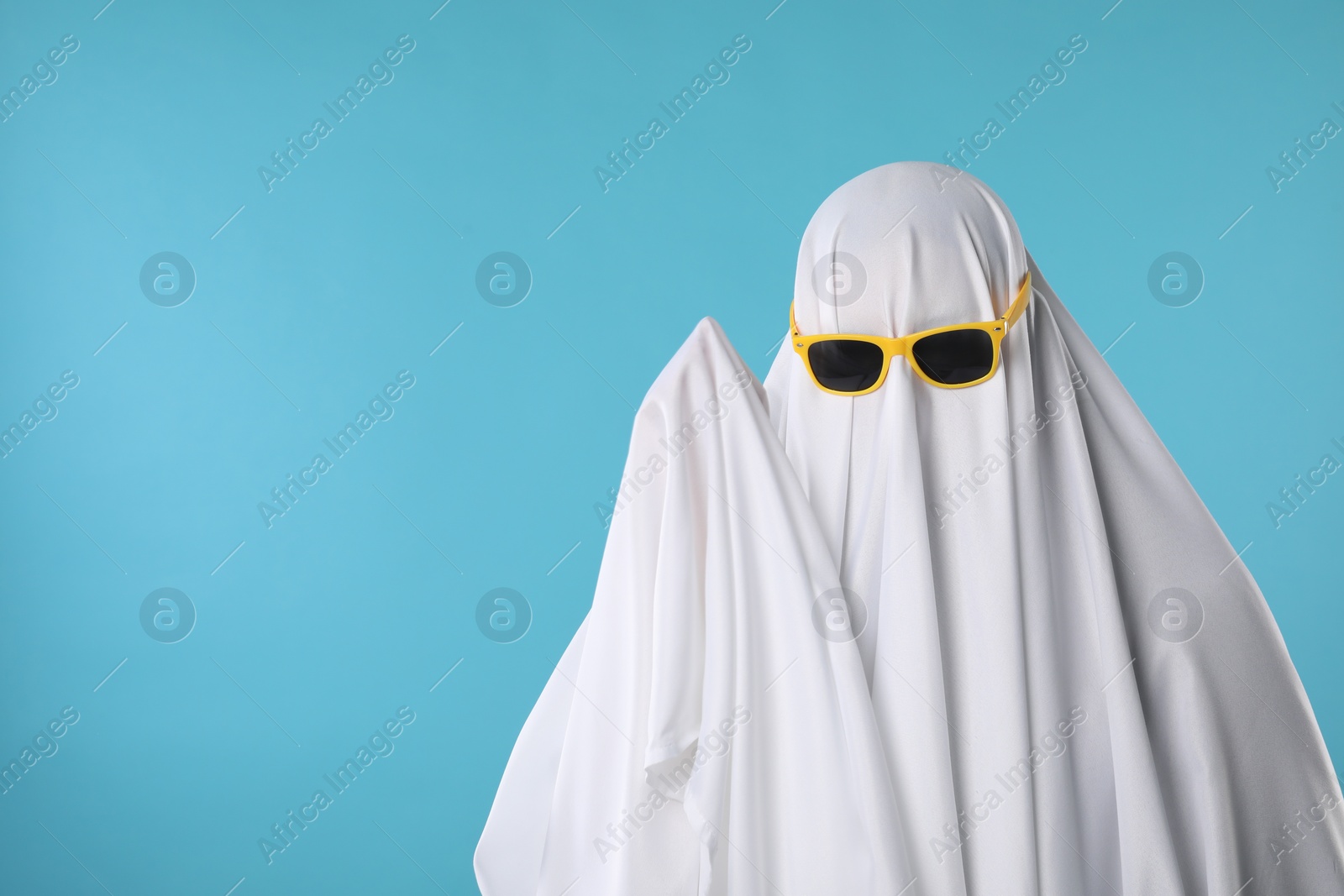 Photo of Stylish ghost. Person covered with white sheet in sunglasses on light blue background, space for text