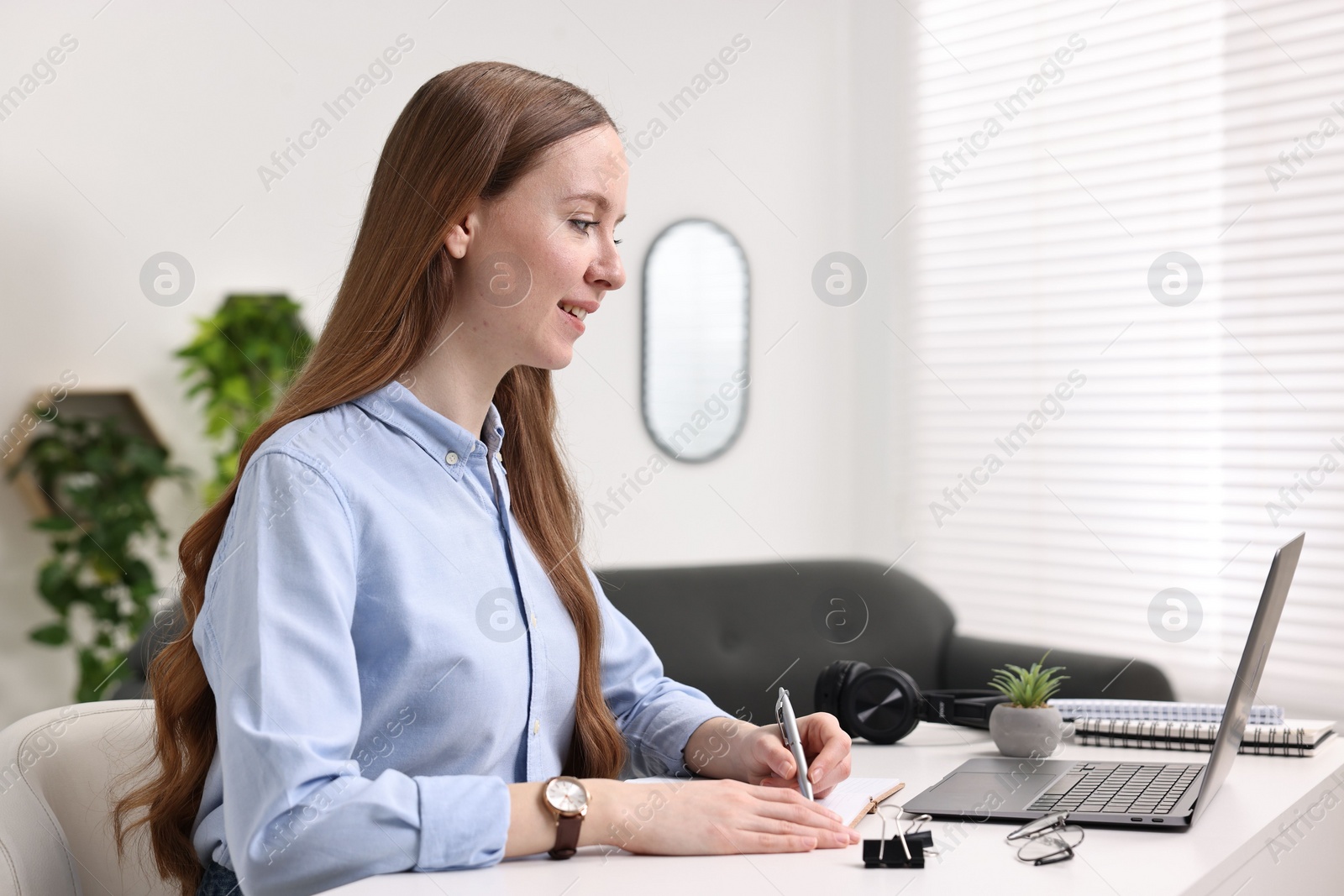 Photo of E-learning. Young woman taking notes during online lesson at white table indoors