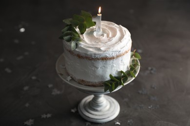 Photo of Tasty Birthday cake with burning candle and eucalyptus branches on grey table