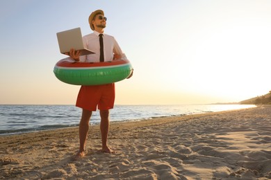 Photo of Happy man with inflatable ring and laptop on beach, space for text. Business trip