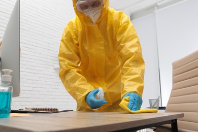 Photo of Janitor in protective suit disinfecting office furniture to prevent spreading of COVID-19