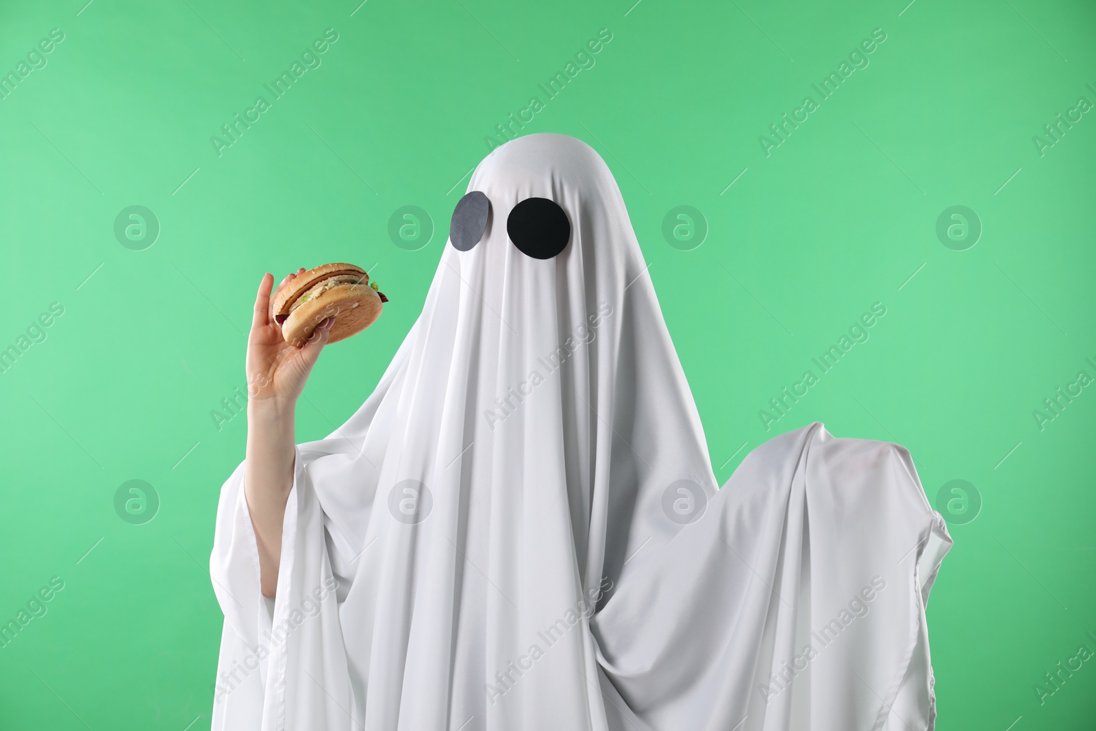 Photo of Creepy ghost. Person in white sheet with tasty hamburger on green background