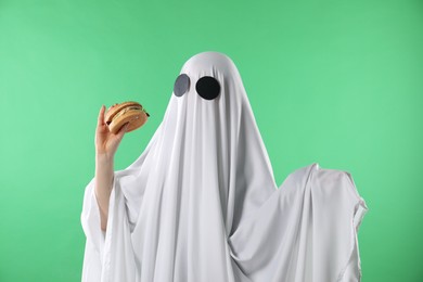 Creepy ghost. Person in white sheet with tasty hamburger on green background